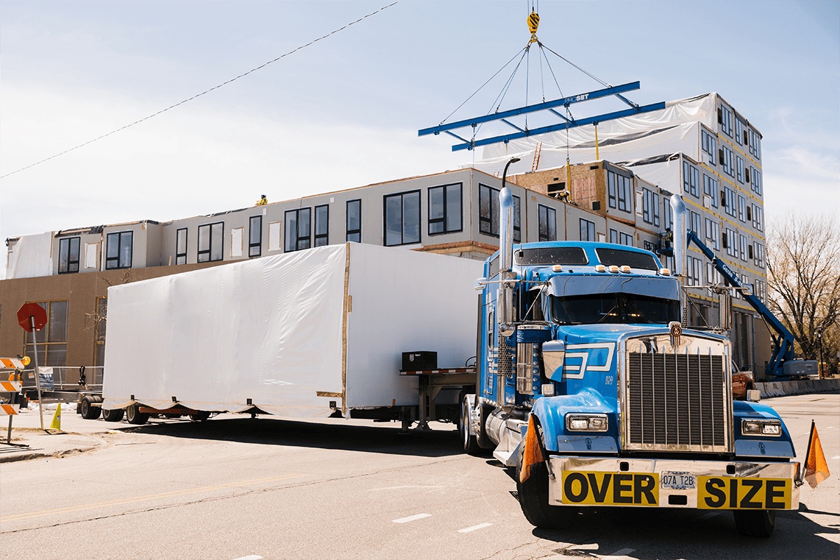 Image of tractor trailer backing into construction site with a modular piece of an apartment building that is under construction.